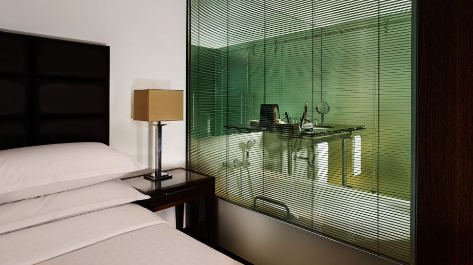 Habitación de hotel con una gran ventana con persiana integrada que deja ver el aseo al otro lado