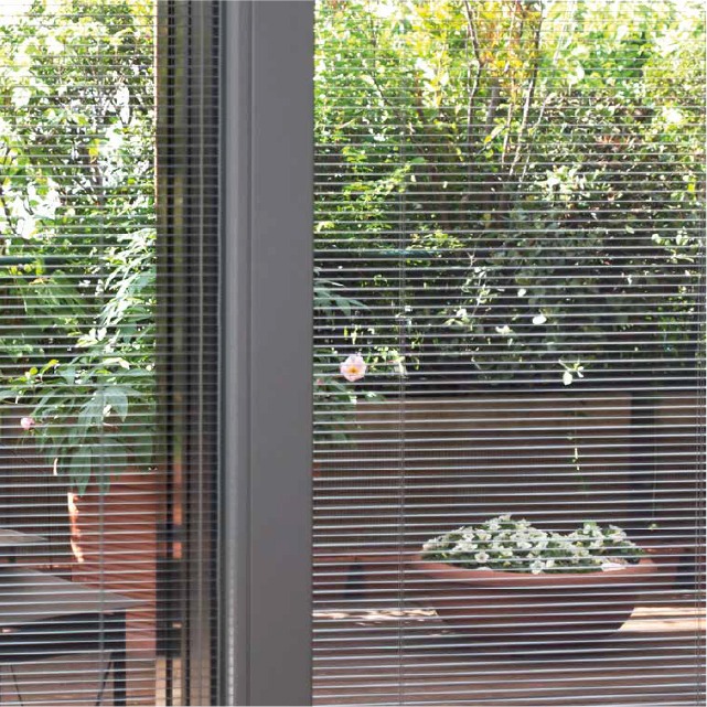 Detalle de una persiana veneciana integrada en una ventana a través de la cual se ve un jardín